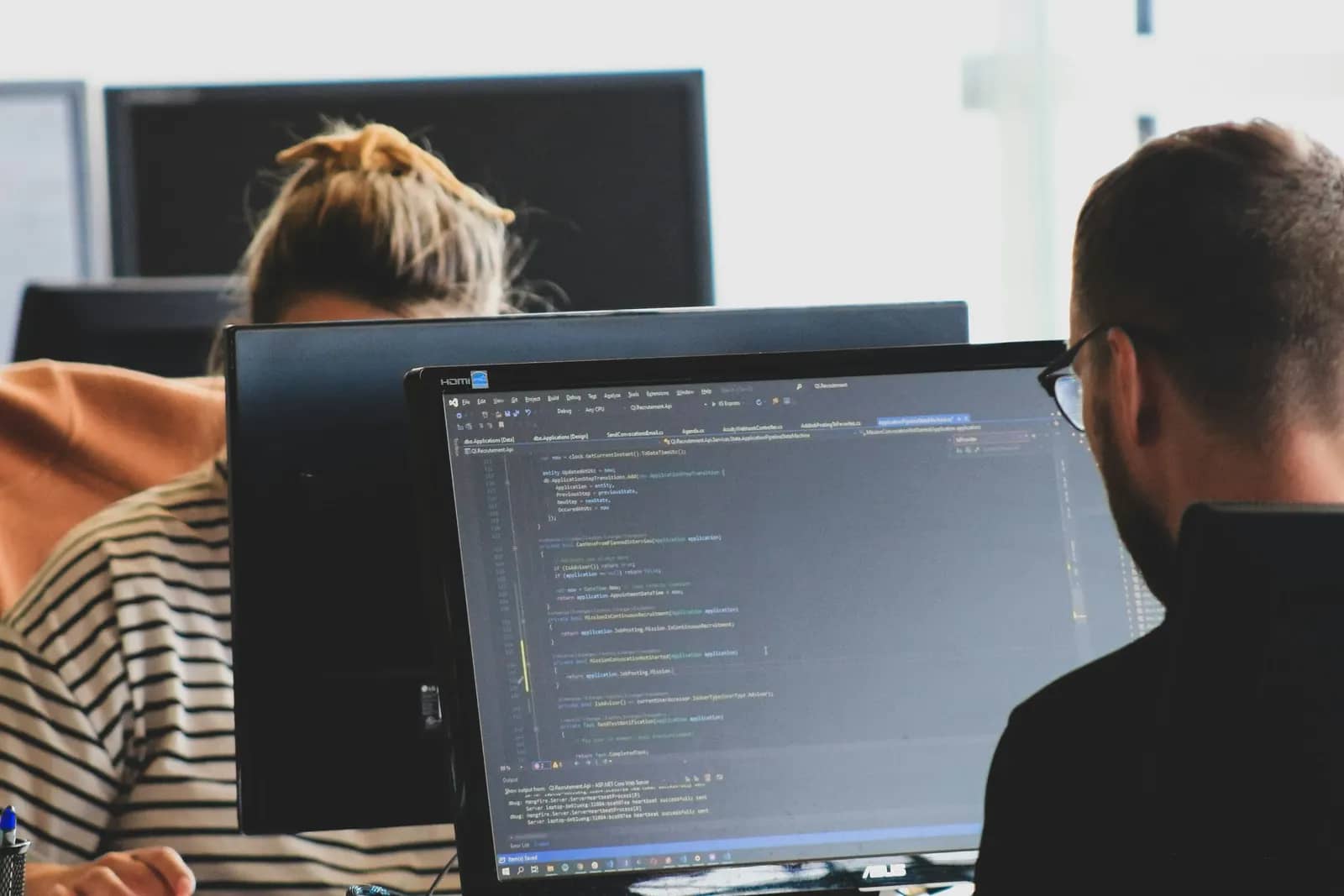 Two engineers working on code across facing monitors in a bright office.
