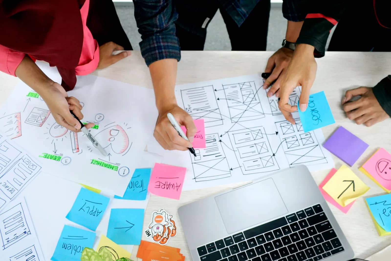 A product team sketching app wireframes on paper beside sticky notes and a laptop.