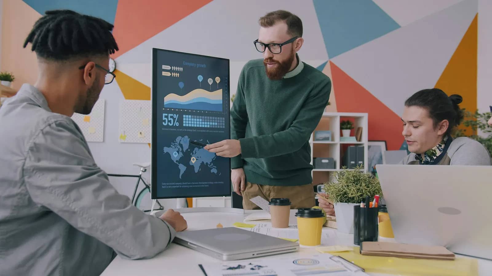 An analyst points to data charts and a world map on a large display while two colleagues follow along.